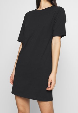 Kvinde iført en ensfarvet sort kortærmet T-shirt-kjole med rund hals, stående mod en ensfarvet lys baggrund.