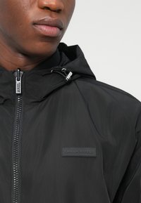 Μαύρο μπουφάν με κουκούλα από ελαφρύ υλικό. Διαθέτει φερμουάρ, ρυθμιζόμενα κορδόνια και μια επώνυμη μπαλώτα στο στήθος.