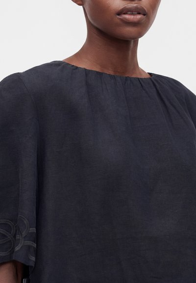 Femme portant un haut ample bleu marine foncé avec des manches trois-quarts et un subtil détail de broderie près de l'ourlet de la manche gauche.