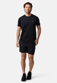 Mustat urheilupaidat lyhyillä hihoilla ja pienellä logolla rinnassa, yhdistettynä mustiin shortsseihin. Mallilla on yllä mustat lenkkarit.