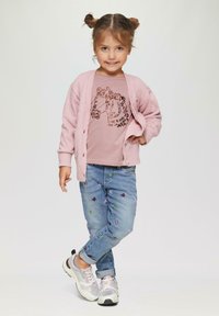 Jasnoróżowy kardigan na różowej koszulce z grafiką, w połączeniu z niebieskimi dżinsami ozdobionymi kolorowymi haftami kwiatowymi. Srebrne buty sportowe dopełniają stylizację.