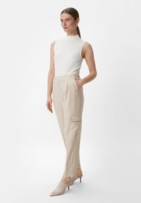 Biała bezrękawnik, beżowe spodnie z plisami i bocznymi kieszeniami oraz plecione czółenka na spiczastym obcasie, na jasnoszarym tle.