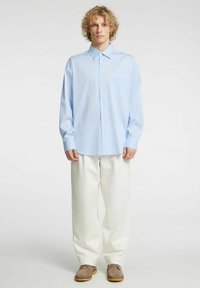 Jasnoniebieska koszula w paski na guziki z kieszenią na piersi, długimi rękawami, luźnym krojem, zestawiona z białymi spodniami i brązowymi butami.