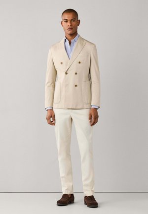Ein Mann steht vor einem schlichten Hintergrund und trägt einen beigen, doppelt geknöpften Blazer, ein hellblau gestreiftes Hemd, weiße Hosen und braune Wildleder-Slipper.
