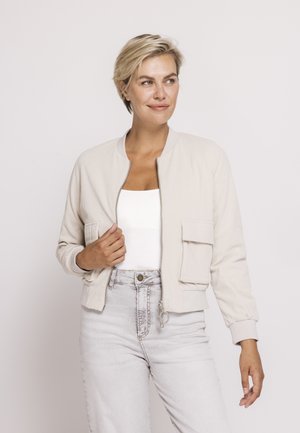 Frau mit kurzen blonden Haaren, die eine hellbeige Jacke mit Reißverschluss über einem weißen Top und hellgrauen Jeans trägt, vor einem schlichten Hintergrund stehend.