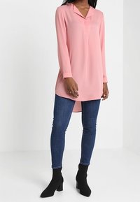 Chemise rose clair à manches longues avec col et patte de boutonnage, associée à un jean skinny bleu foncé et des bottines noires. Tissu lisse, coupe droite.