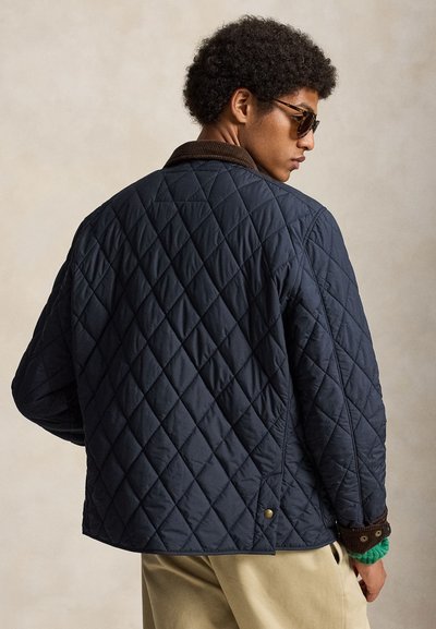 Homme portant une veste matelassée bleu foncé avec un col marron, un pantalon beige, des lunettes de soleil et le bord d’un pull vert, vu de dos sur un fond uni.