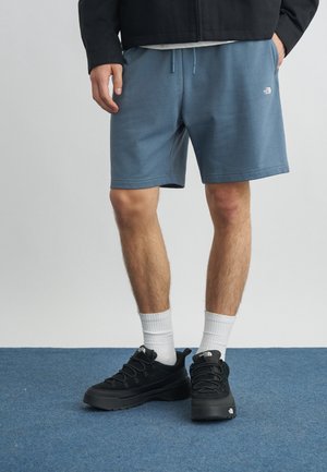 Άτομο με μπλε σορτς με κορδόνια, μαύρα αθλητικά παπούτσια, λευκές κάλτσες και μαύρο μπουφάν που στέκεται σε μπλε χαλί.