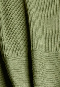 Gros plan sur un tissu vert olive avec un tricot côtelé vertical et une bordure horizontale côtelée à l'ourlet, montrant la texture et les plis.