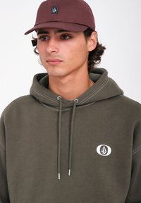 Bruine pet met logo, olijfgroene hoodie met witte stiksels, trekkoorden met metalen tips en een ronde logodetail op de borst.