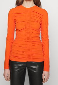 Långärmad orange topp med samlade tyglappar, åtsittande silhuett, rund halsringning och en slät textur, kombinerad med svarta skinnbyxor.