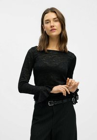Kvinde med skulderlængde brunt hår iført en sort blonde bluse med lange ærmer og sorte stribede bukser, der justerer sin ærme.