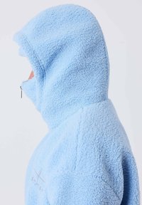 Personne portant une veste à capuche en polaire texturée bleu clair, entièrement fermée par une fermeture éclair, vue de profil sur un fond uni.