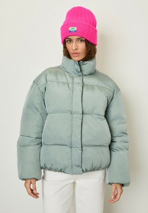 Puffy, light green jacket with a high collar and horizontal stitching. Paired with a bright pink knit beanie featuring a label.
