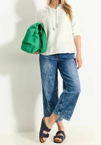 Sudadera blanca con capucha y cordones, vaqueros de pierna ancha azules, sandalias navy y bolsa tote verde. Enfoque en un estilo casual y cómodo.