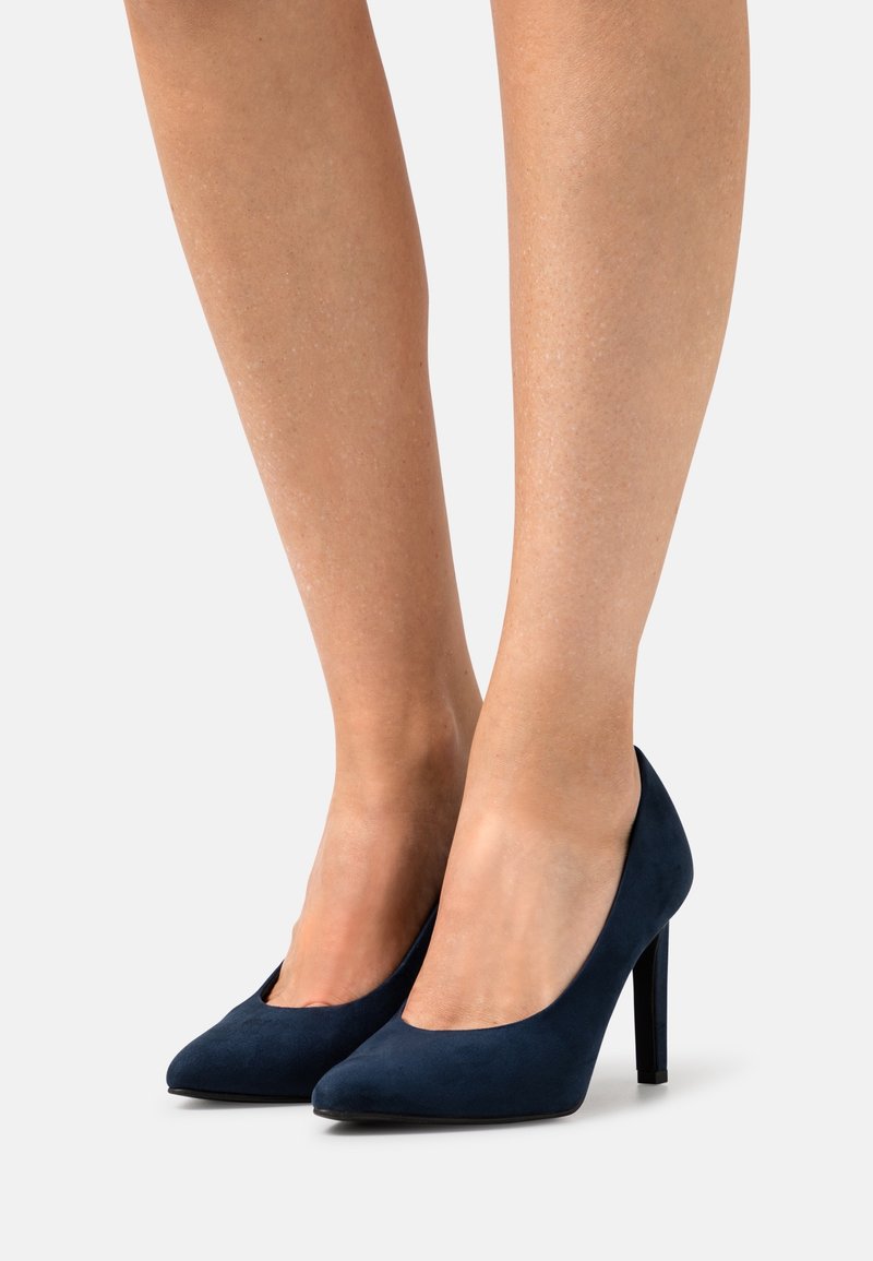 Les jambes inférieures d'une femme portant des escarpins en daim bleu marine à bout pointu avec talons aiguilles sur fond blanc.