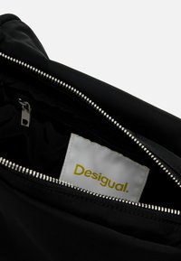 Svart väska med silverdragkedja, med en slät yta och en vit etikett med varumärket "Desigual" i gul text inuti.