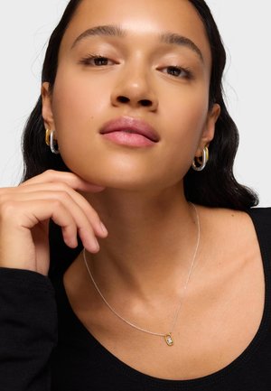 Młoda kobieta o długich ciemnych włosach nosi srebrno-złote kolczyki koła oraz delikatny srebrny naszyjnik z złotą zawieszką.