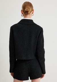 Musta tweed-takki, jossa on kimaltelevia lankoja, lyhyt malli, notched-kollari ja pitkät hihat, yhdistettynä vastaaviin shortseihin. Rakenne on teksturoitu.
