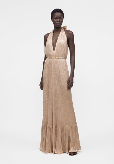 Γυναίκα με μακρύ μέχρι το πάτωμα, αμάνικο μπεζ φόρεμα με βαθύ V-λαιμόκοψη και μικρή ζώνη, που στέκεται μπροστά σε απλό λευκό φόντο.