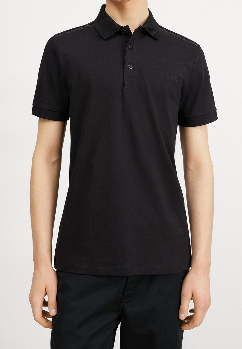 Homme portant un polo noir uni à manches courtes avec un col et une patte à deux boutons, associé à un pantalon noir.