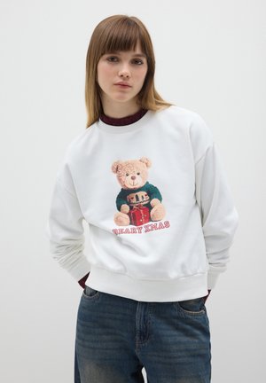 Jeune femme avec des cheveux lisses châtain clair portant un sweat-shirt blanc avec un ours en peluche tenant un cadeau et le texte "BEARY XMAS", associé à un jean bleu.