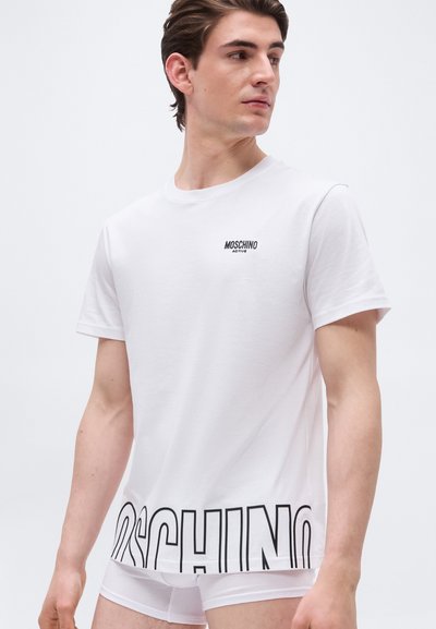 Άνδρας που φοράει λευκό t-shirt Moschino Active με μεγάλο μαύρο λογότυπο στο τελείωμα, κοιτάζοντας προς το πλάι, σε απλό λευκό φόντο.