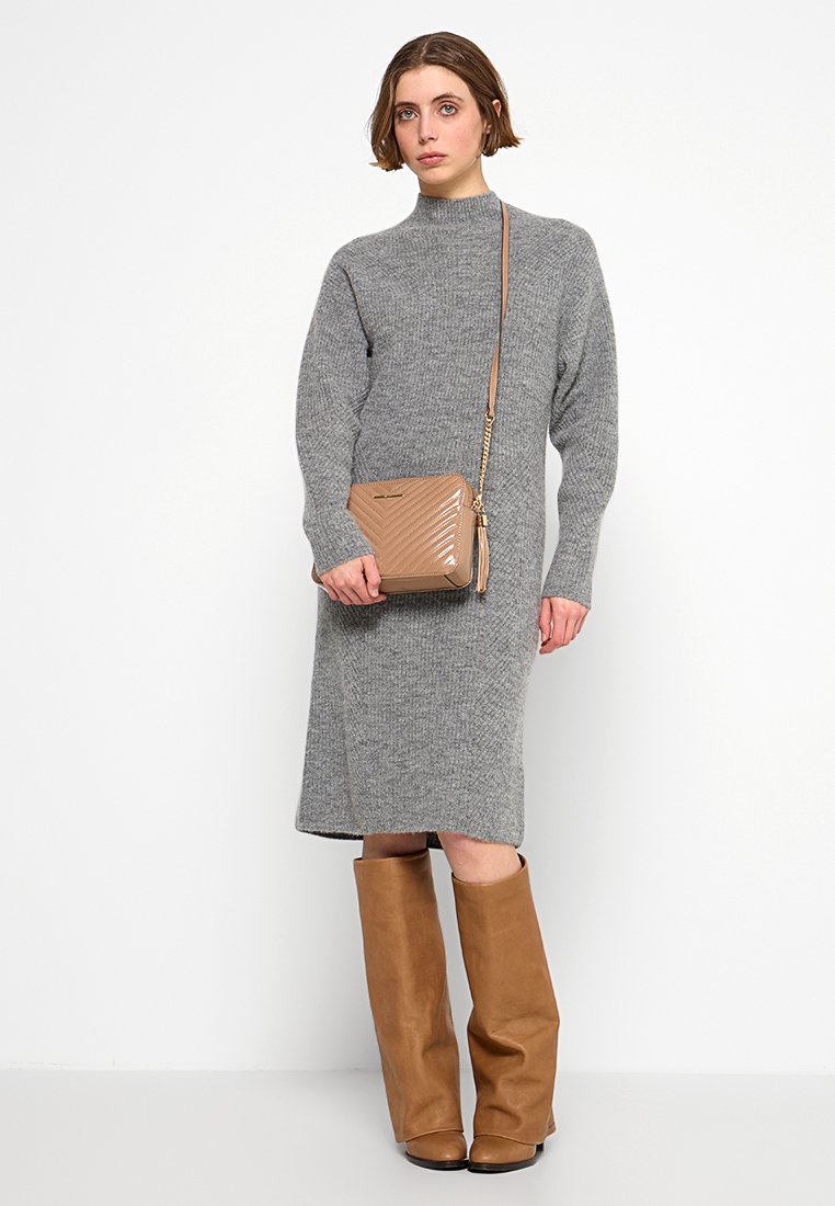 Femme portant une robe midi en tricot gris, des bottes marron jusqu'aux genoux et un sac bandoulière matelassé beige, se tenant devant un fond blanc.