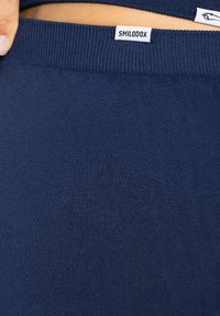 Tissu extensible bleu marine avec une texture côtelée. Comprend une étiquette blanche avec "SMILODOX" au niveau de la taille. Détail en gros plan du matériau.