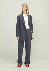 Fato cinza com um blazer ajustado e calças de perna reta, combinado com uma camisola branca curta e sapatos slip-on texturizados vermelhos brilhantes.