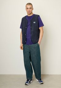 Navy modrá fleecová vesta s gombíkovým zapínaním cez fialové tričko, skombinovaná s voľnými tmavozelenými džínami a šedými teniskami.