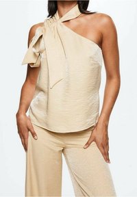 Haut sans manches beige avec un détail de nouage au cou, présentant une texture lisse et un léger éclat, associé à un pantalon large assorti.