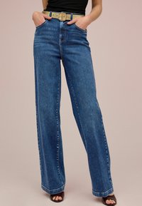 Jean en denim bleu à jambes larges avec une taille haute, présentant des poches avant et une ceinture à boucle dorée. Texture lisse avec des détails de couture classiques.
