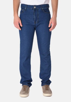 Uomo che indossa jeans blu di denim e sneakers beige, in piedi davanti a uno sfondo chiaro uniforme con le mani rilassate ai lati.