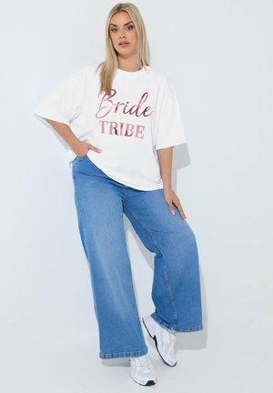 Blond kvinde iført en hvid oversized T-shirt med teksten "Bride Tribe", vide blå jeans og hvide sneakers foran en ensfarvet lys baggrund.
