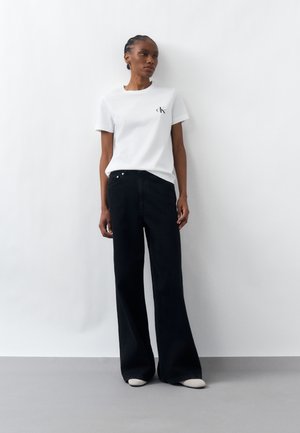 Kobieta ubrana w białą koszulkę z logo CK i szerokie czarne jeansy, stojąca na tle białej ściany z szarą podłogą.