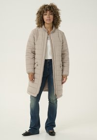 Manteau long beige rembourré avec une fermeture éclair à l'avant, un col côtelé et deux poches latérales. Porté par-dessus un haut blanc et un jean évasé en denim.