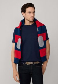 Camiseta navy con bolsillo, llevada debajo de un sudadera con capucha de rayas que presenta rayas rojas y azules, con acentos grises en las mangas.