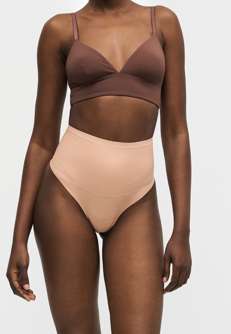 Γυναίκα που φορά καφέ bralette και μπεζ εσώρουχο χωρίς ραφές με ψηλή μέση, σε απλό ανοιχτό φόντο.