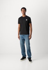 T-shirt en coton noir à manches courtes avec un petit graphique sur la poitrine gauche, associé à un jean en denim bleu clair et des chaussures noires.