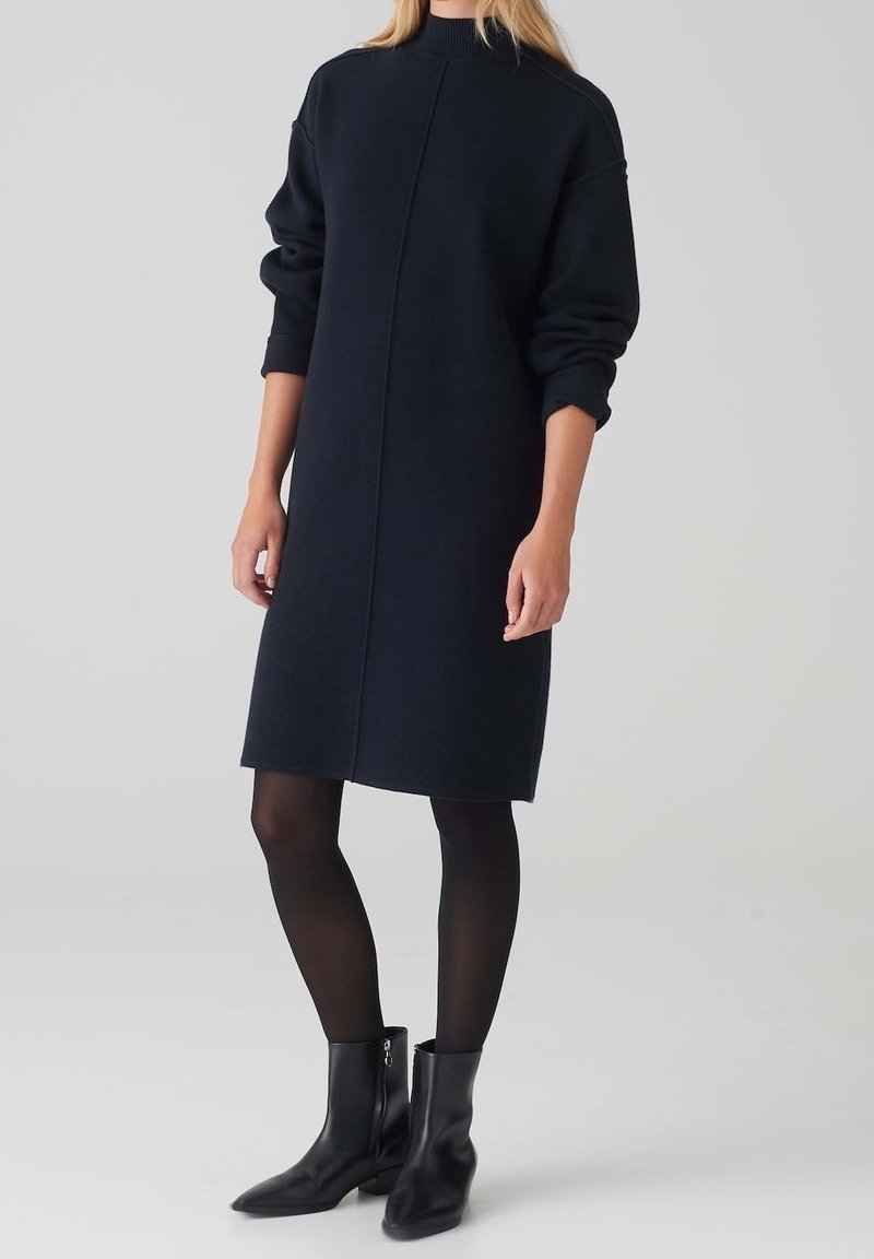 Femme portant une robe en maille noire à manches longues avec un col montant, des collants noirs et des bottines noires, se tenant devant un fond uni.