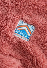 Blødt, lyserødt, fluffy stof med et syet blåt, hvidt og orange rektangulært Superdry Mountain Sport-mærke.