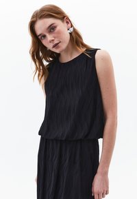 Schwarzes Kleid mit plissierter Textur, ärmellosem Design und rundem Ausschnitt. Das Model trägt silberne Ohrringe in Schmetterlingsform.