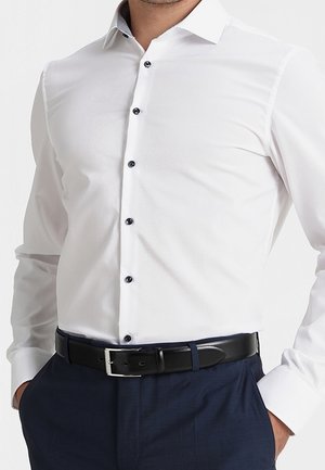 Man draagt een nauwsluitend wit overhemd met zwarte knopen, ingestopt in marineblauwe broek met een zwarte leren riem.