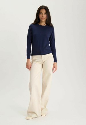 Femme debout portant un pull marine à manches longues et un pantalon large crème sur un fond clair uni.