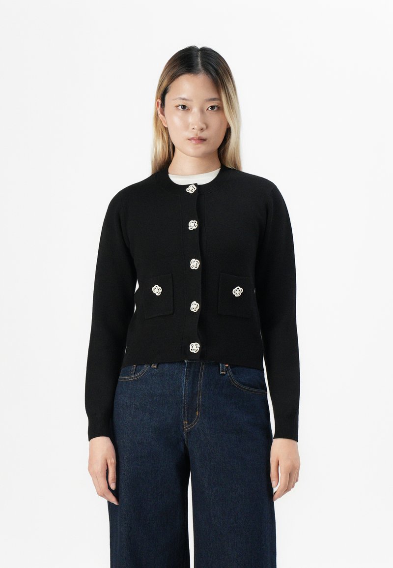 Jeune femme avec des cheveux raides portant un cardigan noir boutonné avec des boutons floraux et un jean large bleu foncé, se tenant devant un fond blanc.
