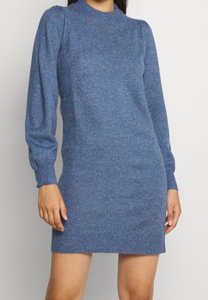 Femme portant une robe-pull bleue ajustée en maille avec de longues manches bouffantes et des poignets et ourlets côtelés.