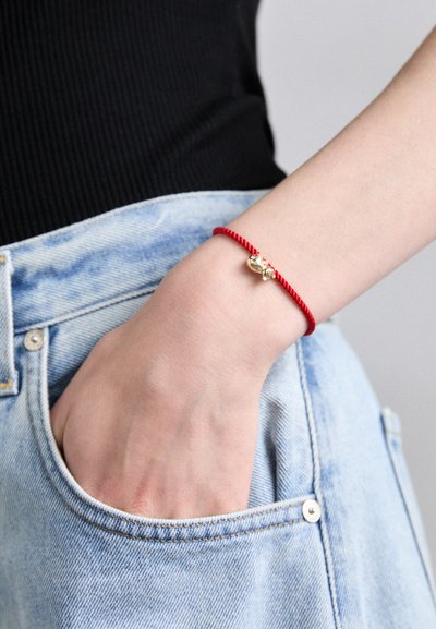Pulsera de cordón rojo retorcido con un charm dorado y acentos transparentes, llevada en una mano que descansa cerca de un bolsillo de mezclilla.