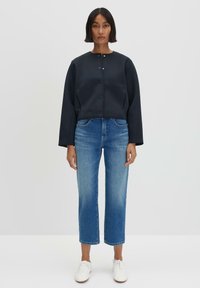Marineblauwe cropped jack met een ronde hals en drukknoopsluitingen, gecombineerd met blauwe rechte spijkerbroeken en witte sneakers. Soepele textuur, casual ontwerp.