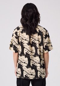 Short-sleeve shirt in beige and black, featuring a swirling abstract face pattern. Made of lightweight fabric with a relaxed fit.